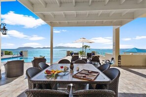 A covered outdoor dining area with wicker furniture, a table set with fruits, wine, and a vase, overlooking a pool and the sea. - Beachfront Bliss: Private Retreat With Spectacular Views (Blowing Point)