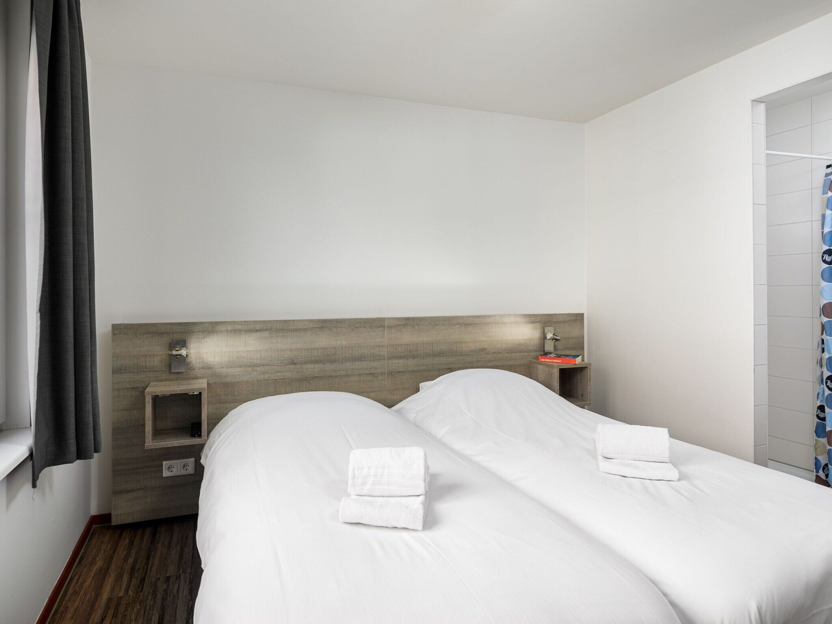A hotel room with two beds, a wooden headboard, a nightstand with a lamp, and a window with curtains.