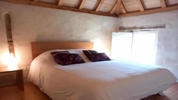 Quarto com piso de madeira, cama com roupa de cama branca e almofadas estampadas, mesa de cabeceira e janela com iluminação natural.