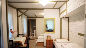 A small, well-lit room with a wooden floor, a single bed, a small table with a vase of flowers, a mirror on the wall, and a door leading to another room.