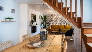 Une table de salle à manger en bois, agrémentée d’un vase et d’un saladier aux motifs géométriques, un canapé jaune et un escalier avec des rampes en bois.