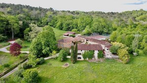 Un grand bâtiment historique doté de nombreuses pièces et d’un court de tennis, entouré d’une végétation luxuriante et d’une colline boisée.