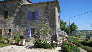 Una casa de pedra amb un camí de grava, una barbacoa i un jardí ple de flors i arbustos.