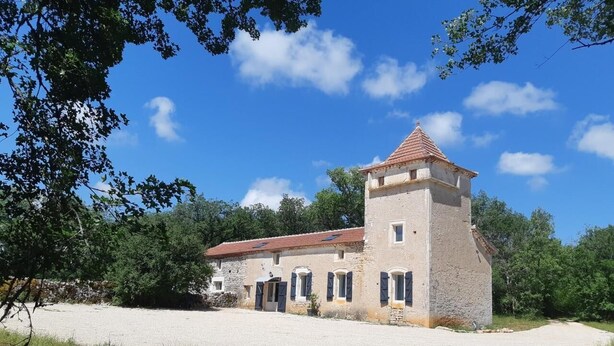 Ein Gebäude aus Stein mit rotem Ziegeldach und einem kleinen Turm, umgeben von Bäumen und einem Kiesweg.