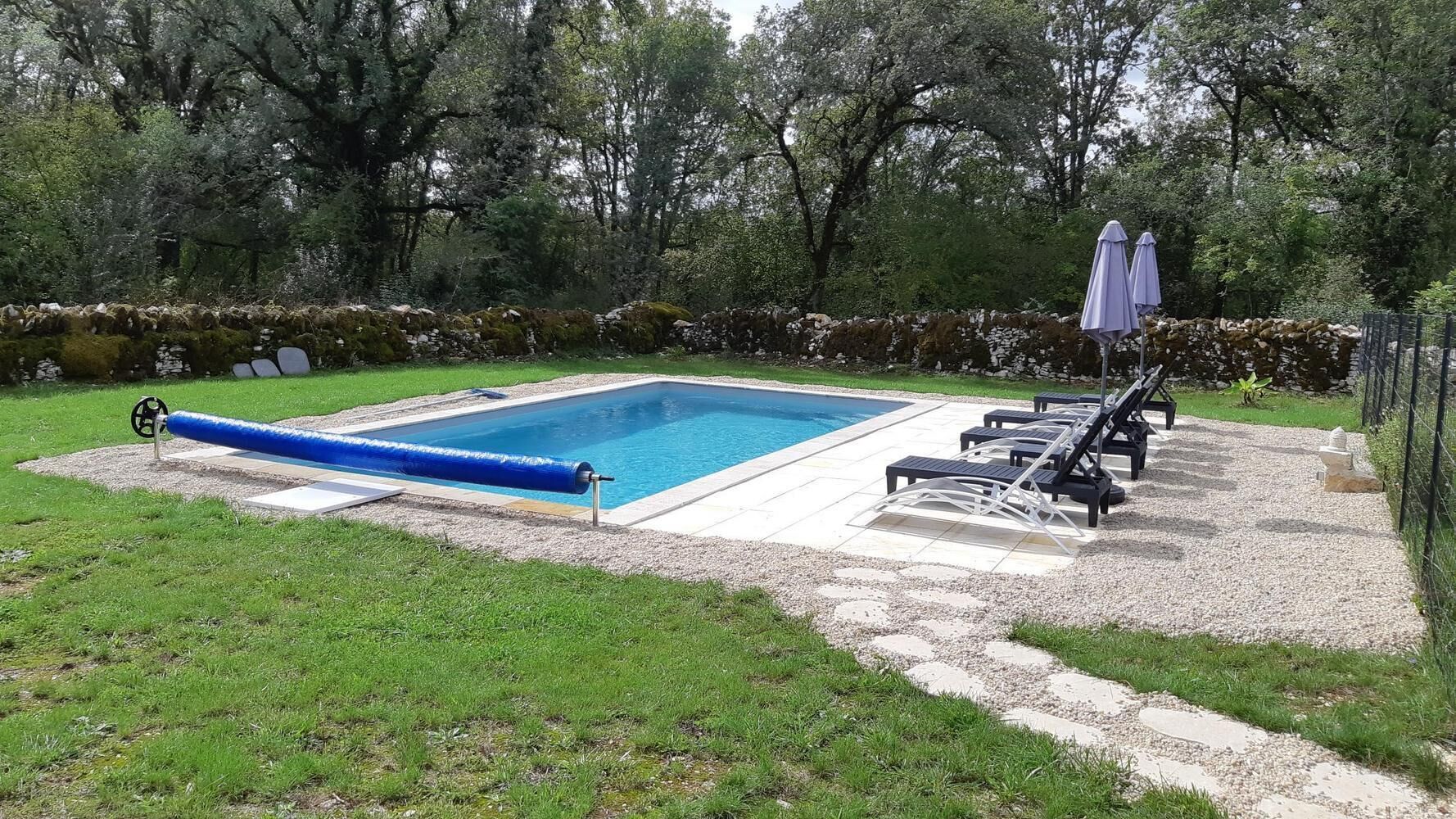 Ein rechteckiger Swimmingpool mit einer blauen Rutsche, umgeben von einer Steinmauer und einer Schotterfläche. Im Hintergrund sind Liegestühle und Sonnenschirme zu sehen.