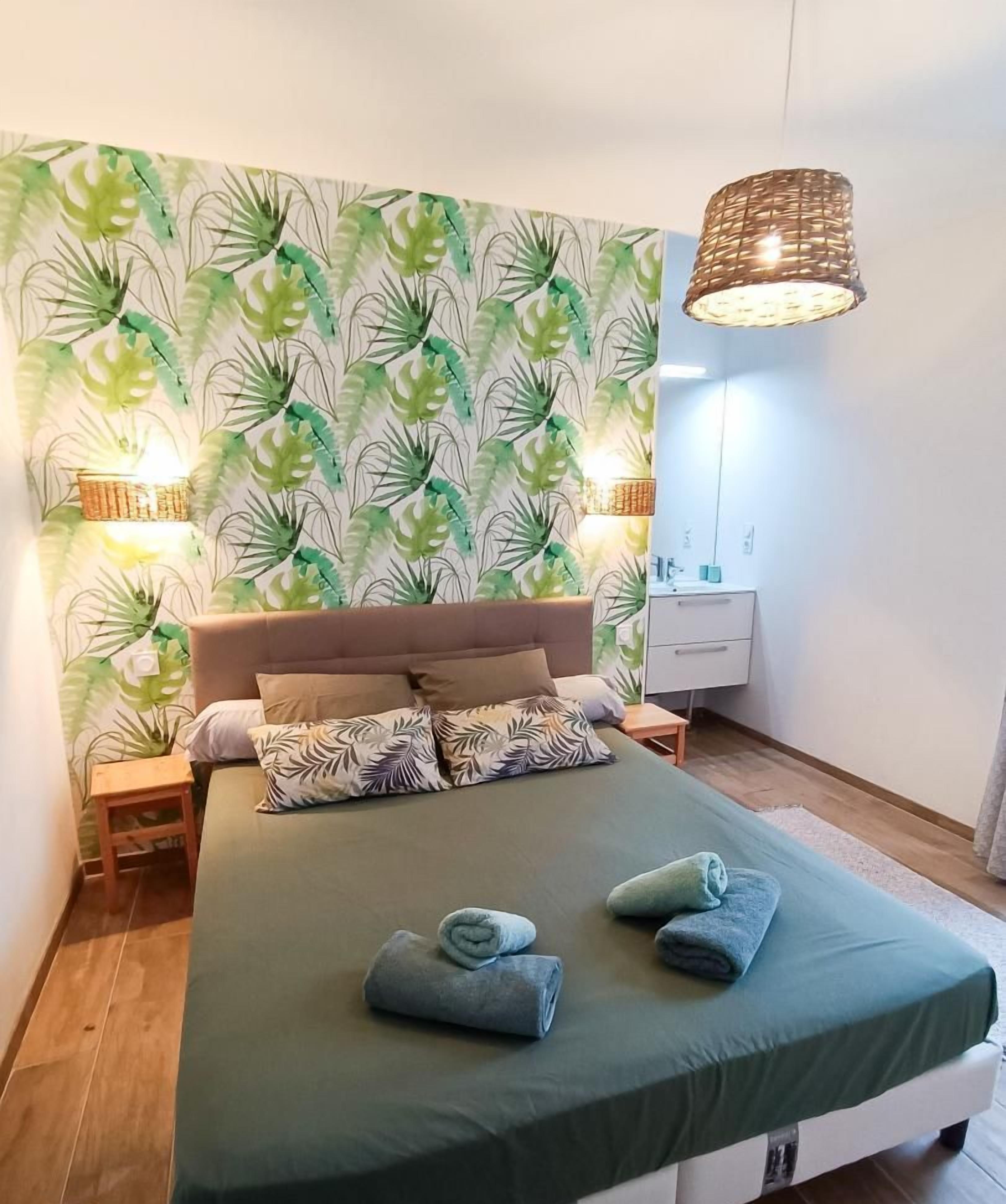A bedroom with a bed, a nightstand, a bedside table, a wall-mounted lamp, and a wall with a leafy wallpaper pattern.