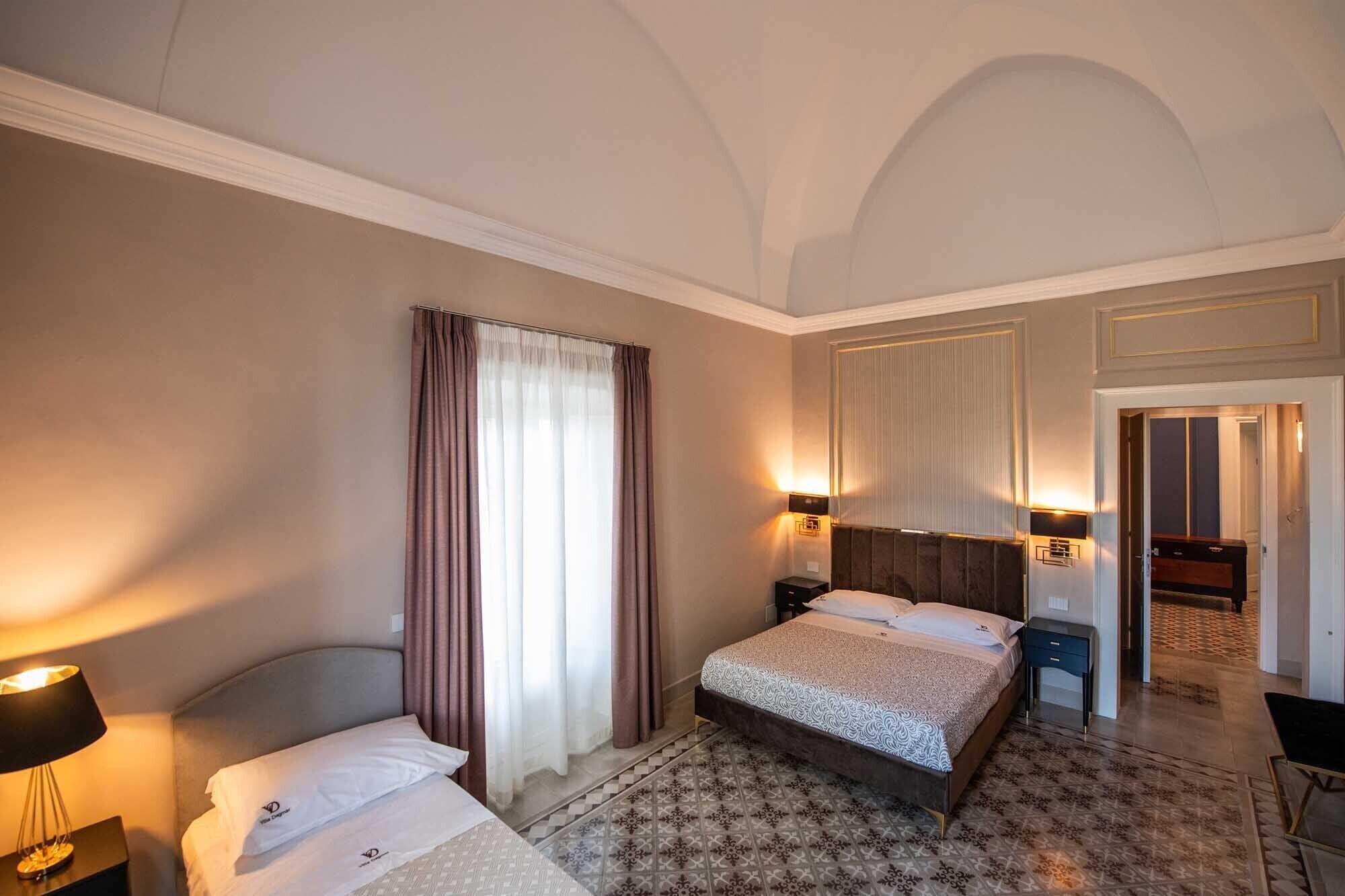 Une chambre d’hôtel comprenant un lit, une table de chevet, une lampe, une fenêtre avec des rideaux et un luminaire fixé au mur.