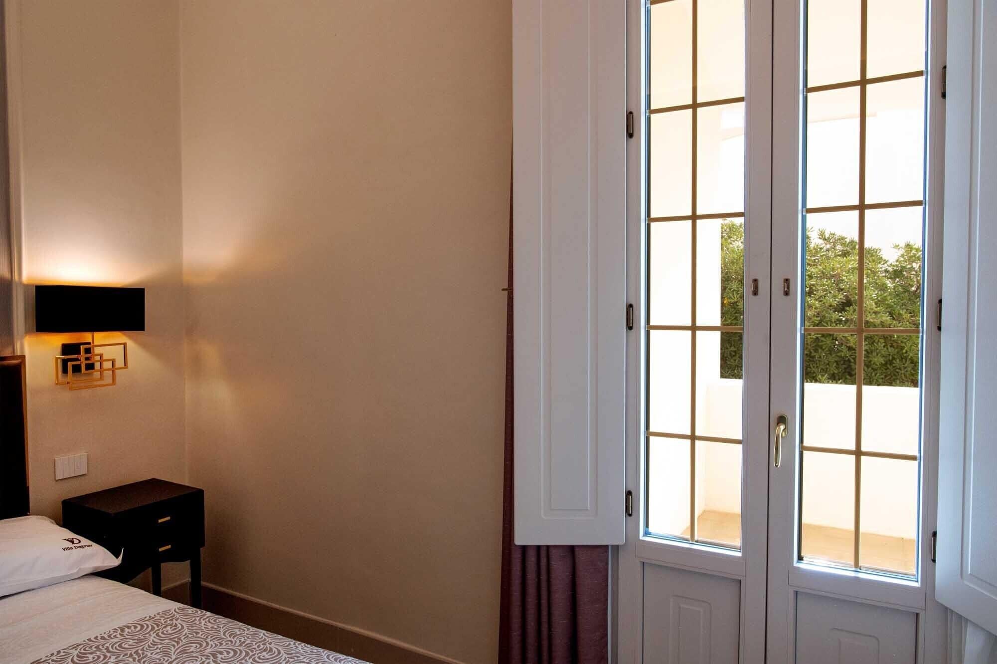 A hotel room with a bed, a bedside lamp, a nightstand, and a large window with a view of greenery outside.