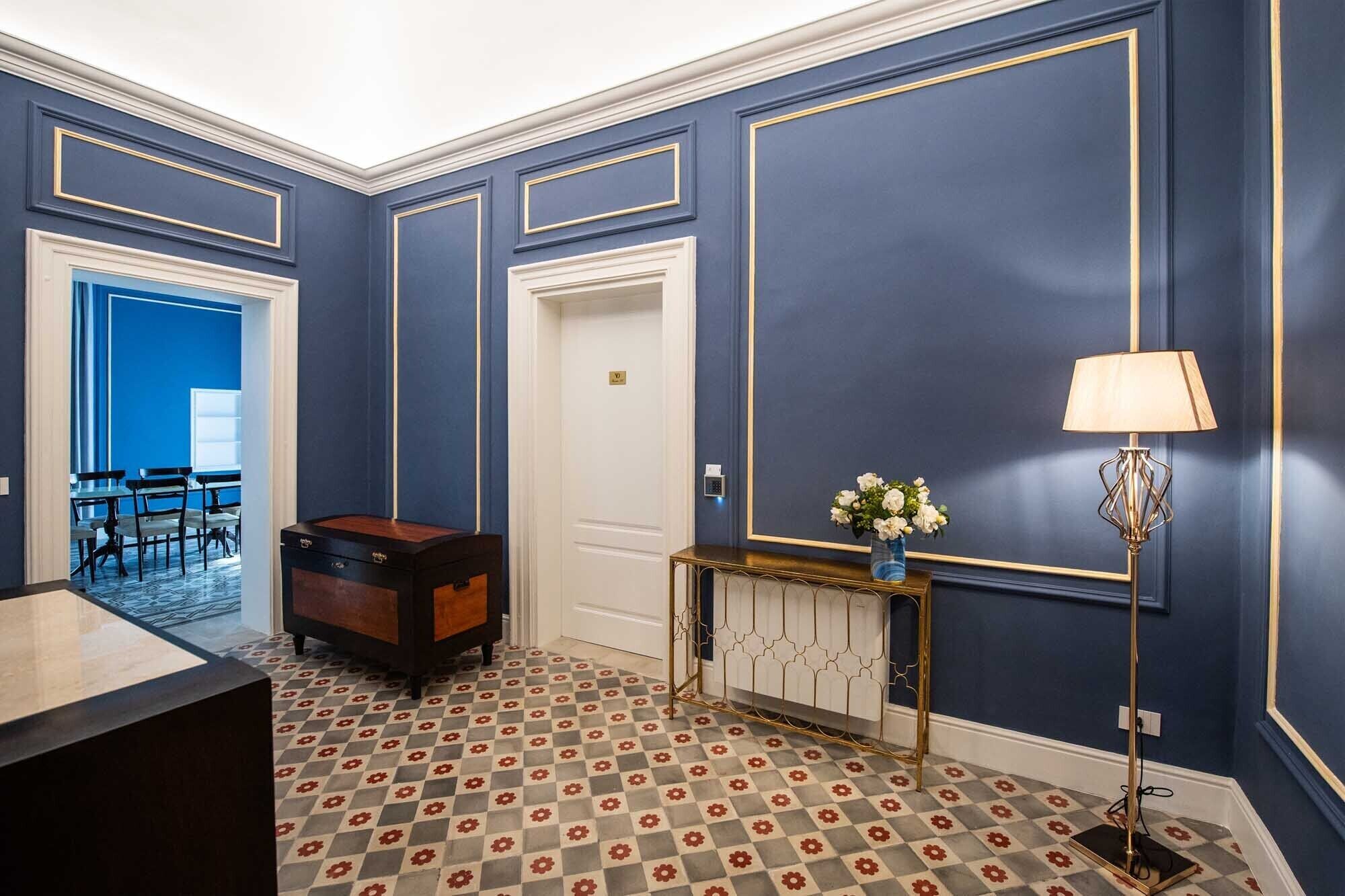A hotel room with a checkered floor, a desk, a chair, a small table with a vase, and a lamp.