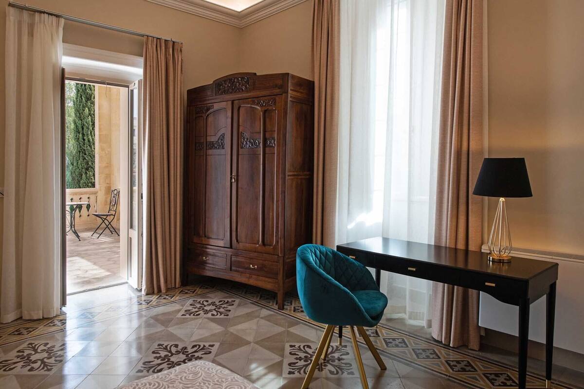 Ein Zimmer mit einem Holzschrank, einem türkisfarbenen Sessel, einem Schreibtisch mit Lampe und einem Boden aus Fliesen mit Muster.