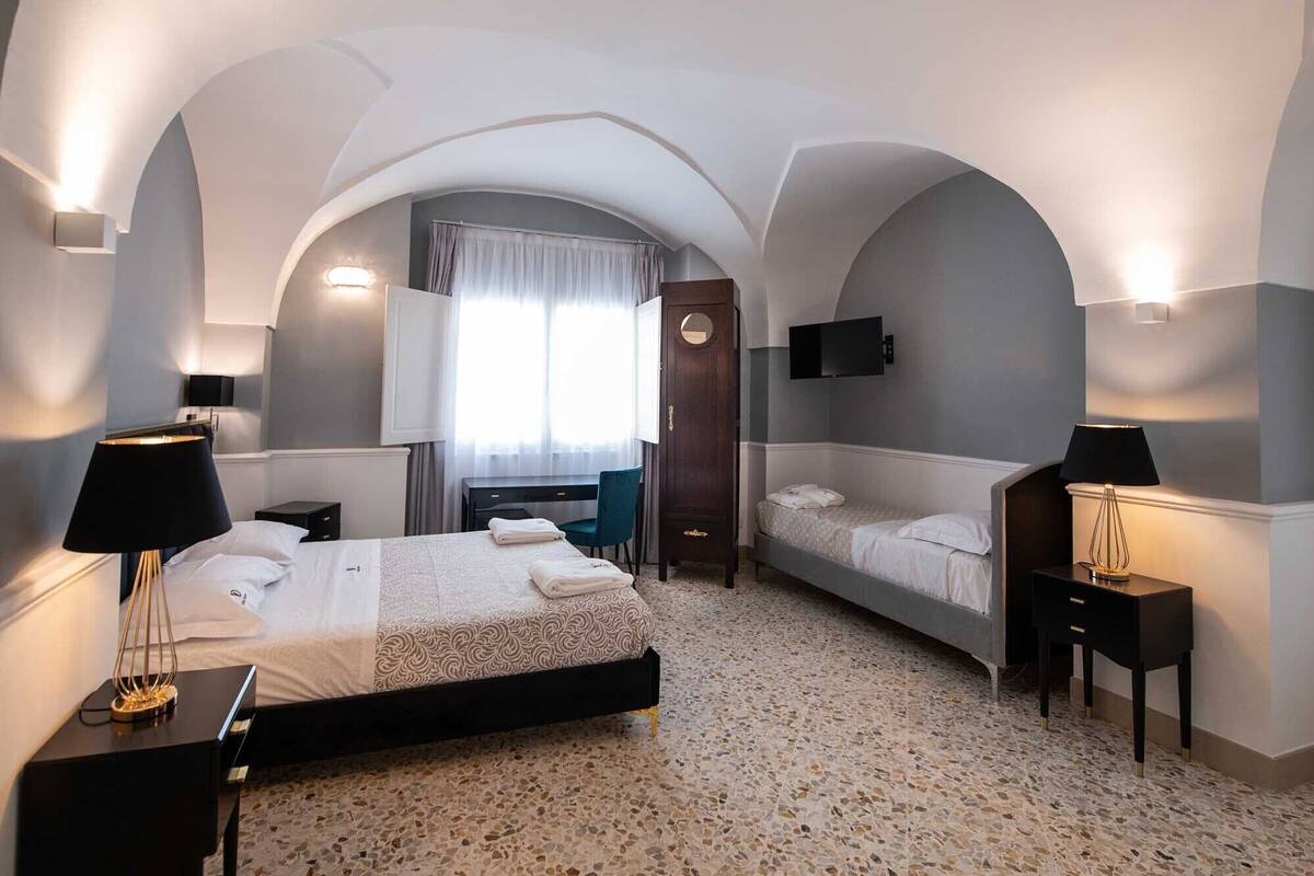 Ein Hotelzimmer mit einem großen Bett, einem Einzelbett, einem Schreibtisch, einem Stuhl, einem Kleiderschrank und einem Fernseher.