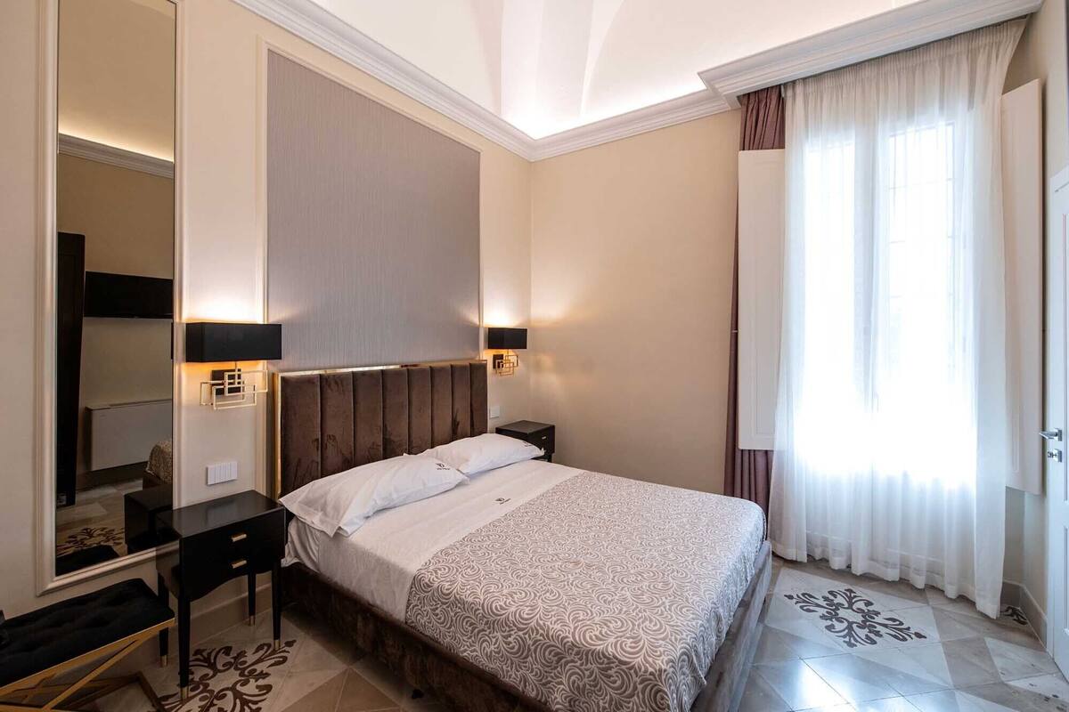 Ein Hotelzimmer mit Bett, Nachttischen, einem Spiegel und einem großen Fenster mit Vorhängen.
