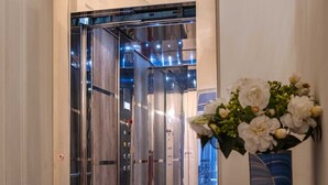 Un ascensor moderno con puerta de vidrio, un florero con flores blancas sobre una consola negra y un suelo de baldosas.