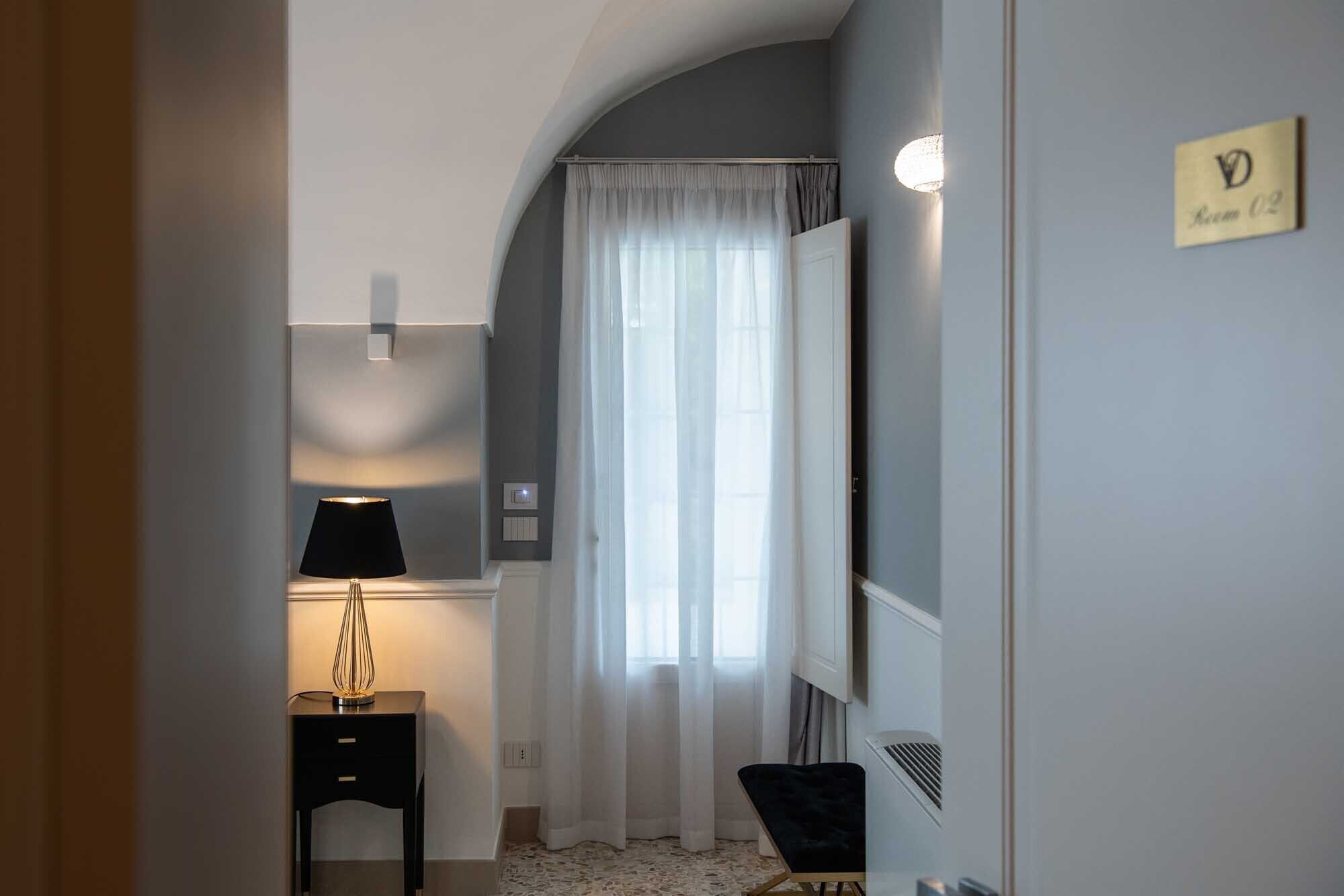 Ein Hotelzimmer mit Bett, Nachttisch mit Lampe, Fenster mit Vorhängen und einem Schild mit der Zimmernummer.