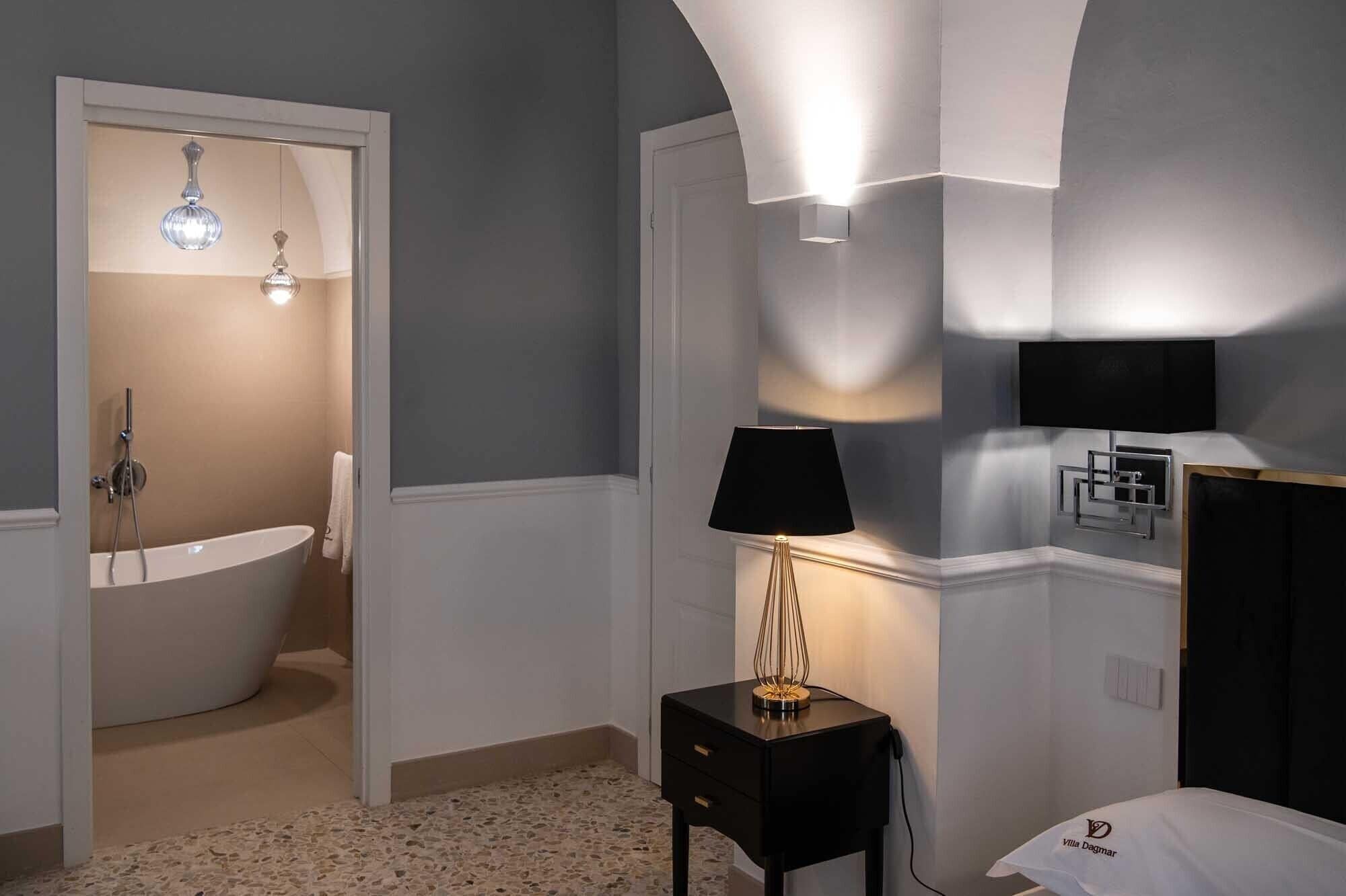 A modern bathroom with a bathtub, a standing shower, a vanity with a mirror, and a bedside table with a lamp.
