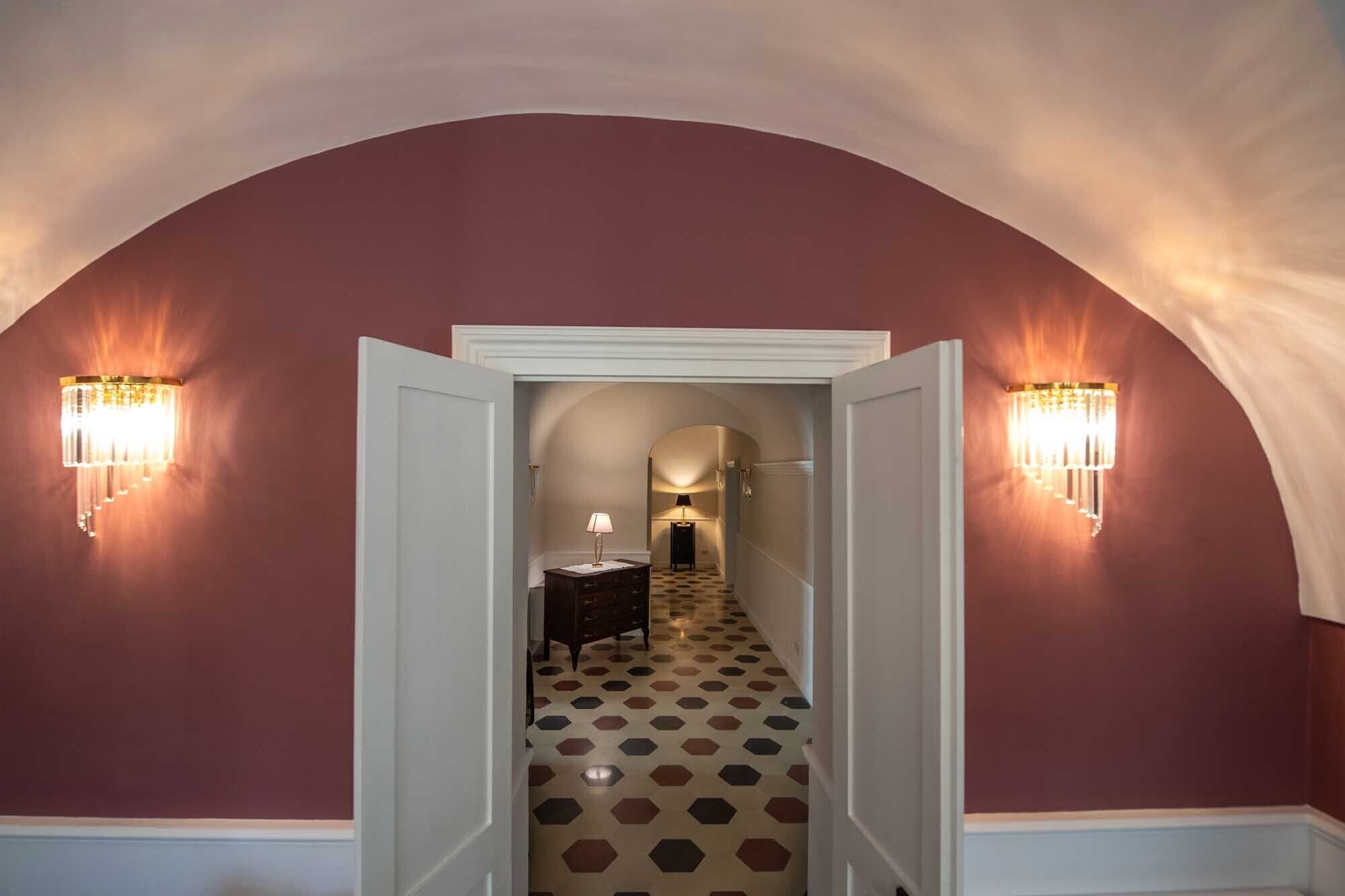 A hallway with a checkered floor, a white door at the end, and wall sconces on either side.
