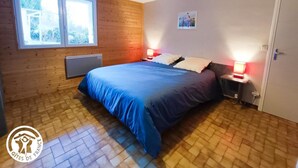 Une chambre avec un plancher en bois, un lit avec une literie bleue, deux tables de chevet avec des lampes, une fenêtre et une porte.