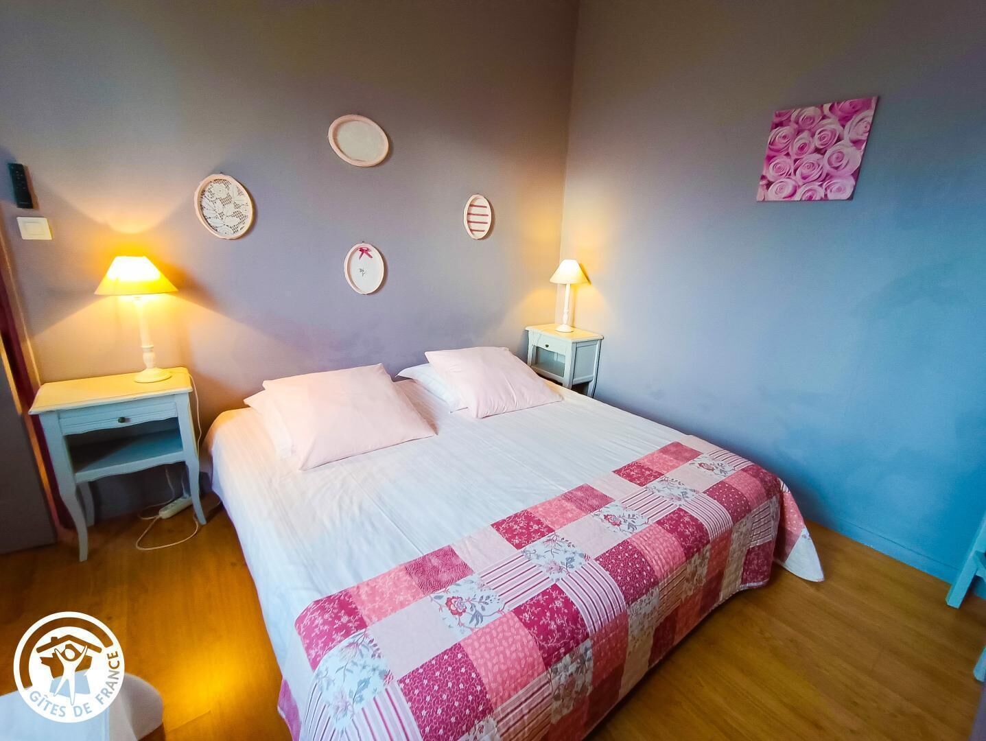 A bedroom with a bed, two bedside tables with lamps, a wall clock, and a floral artwork.