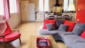 Una sala de estar acogedora con un sofá gris, cojines rojos, una mesa de centro de madera y una butaca roja. Se ve una cocina al fondo con un refrigerador blanco y una estufa.