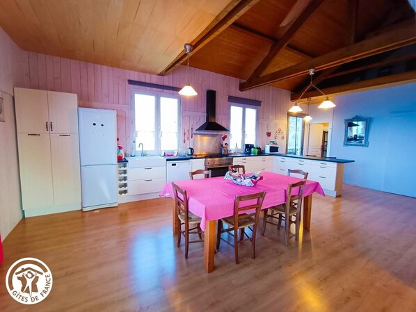 A spacious kitchen with wooden cabinets, a dining table with pink tablecloth, and a large window providing natural light.