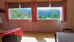 Una habitación con un ventanal grande, dos cortinas a cuadros, un ventilador y una mesa con mantel.