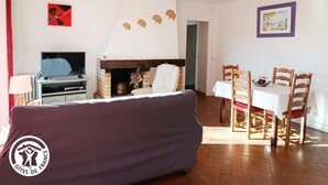 A living room with a sofa, a fireplace, a dining table with chairs, a TV, and artwork on the walls. - Gîtes de France® - La Grange de Marie (RUDELLE)
