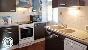 A modern kitchen with dark cabinets, a white stove, a dishwasher, and a window with a plant on the counter. - Gîtes de France® - La Grange de Marie (RUDELLE)