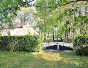 A gated entrance with a hedge, a building with a tiled roof, and trees in the background.