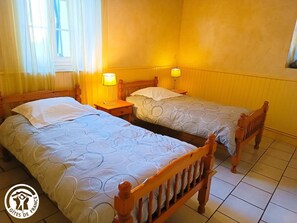 Ein Zimmer mit zwei Holzbettgestellen, jeweils ausgestattet mit einem Nachttisch und einer Lampe. Der Boden ist mit Fliesen belegt und die Wände sind gelb.