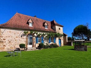 Un bâtiment en pierre avec un toit recouvert de tuiles rouges, une porte d’entrée et un petit porche doté de chaises en métal.