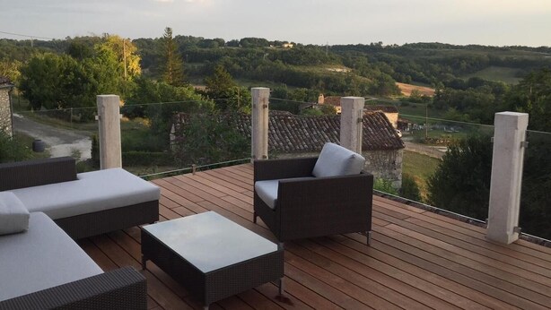 Una terraza de madera con mobiliario exterior, incluyendo sofás y una mesa, con vistas a un paisaje pintoresco con árboles y edificios.