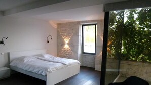 Un dormitorio con una cama, una silla, un ventanal y un muro de piedra.