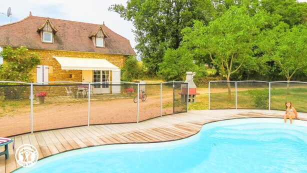 Uma piscina com deck de madeira, uma casa de pedra com toldo branco e um espaço de pátio cercado.