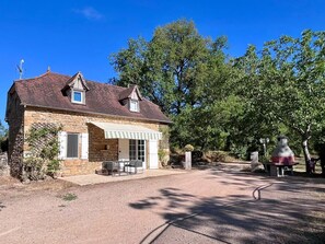 A stone house with a covered patio, a gravel driveway, and a large tree in the background.