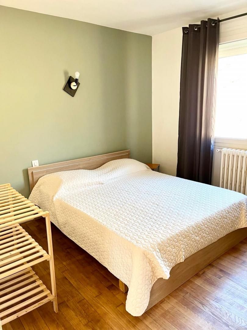 Une chambre à coucher avec un lit, une table de chevet en bois, une lampe fixée au mur et une fenêtre avec des rideaux.