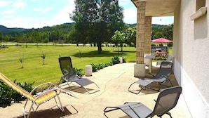A patio with lounge chairs and a table, overlooking a grassy area with trees and a distant hill. - Gîtes de France® - Chez Septi (FIGEAC)