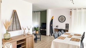 A modern living room with a dining area, featuring a wooden cabinet, a table with a white tablecloth, and a large clock on the wall. - Gîtes de France® - Chez Septi (FIGEAC)