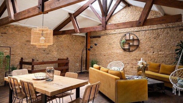 A rustic interior with exposed wooden beams, a stone wall, a wooden dining table with wicker chairs, a yellow sofa, and a hanging wicker light fixture. - Gîtes de France® - Manon (CASSAGNES)