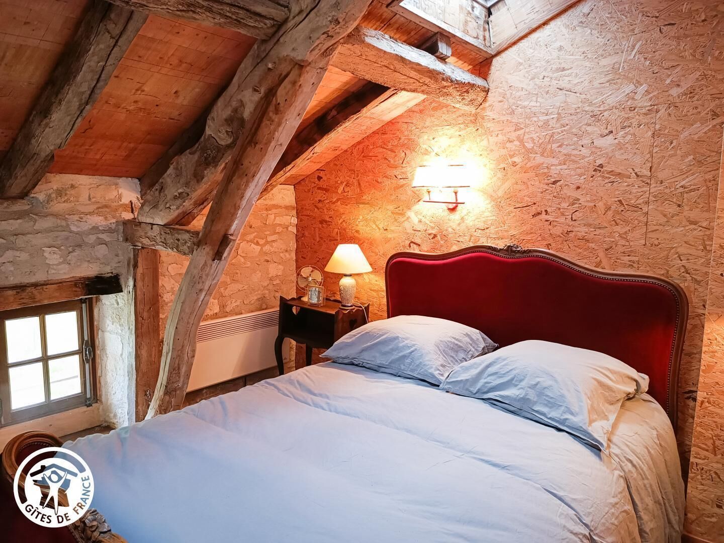 Une chambre avec une tête de lit rouge, un plafond en bois, un mur en pierre et une fenêtre.