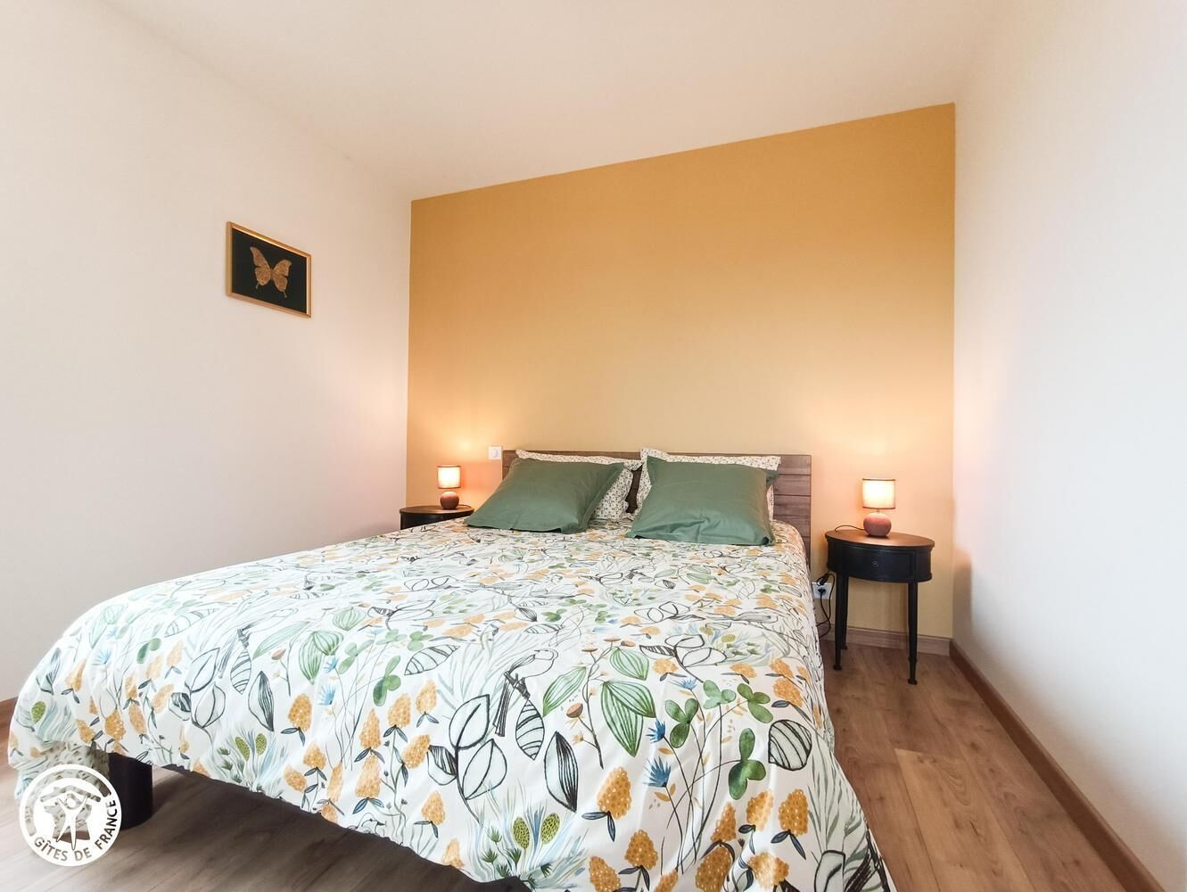 A bedroom with a floral bedspread, two green pillows, a wooden headboard, and two bedside lamps.