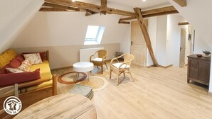 A loft-style room with exposed wooden beams, a sofa with cushions, two wicker chairs, a small round table, and a view of the outside through a skylight.