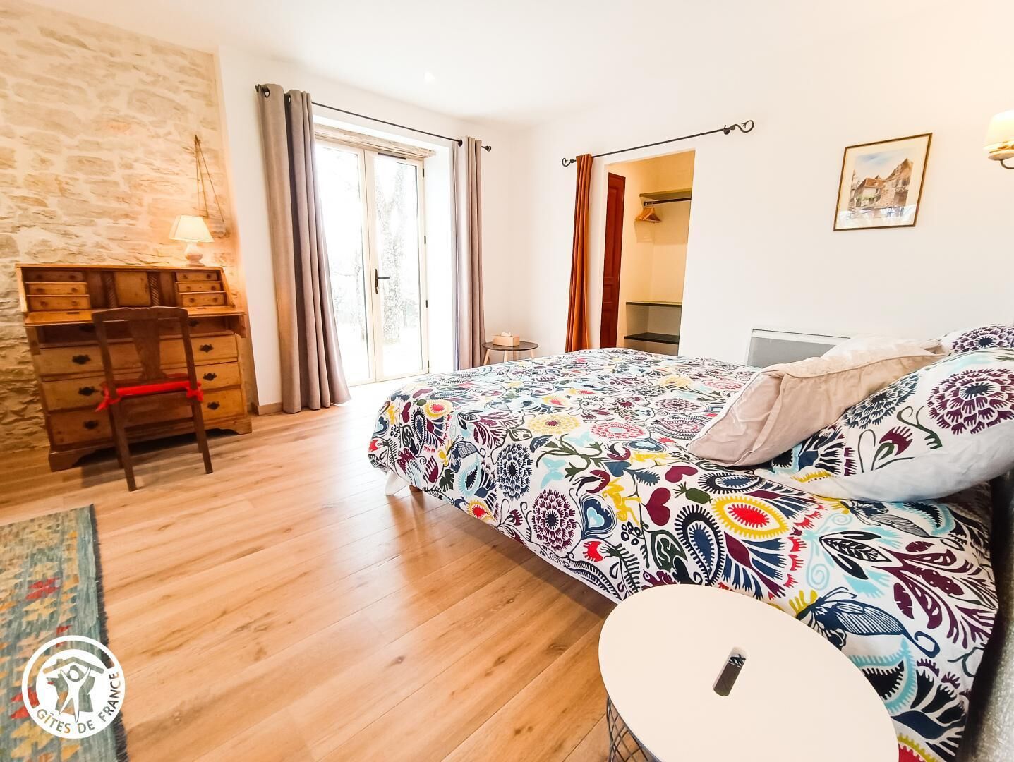 A bedroom with a wooden desk, a bed with a colorful patterned bedspread, a small round table, and a view of the outside through a window.