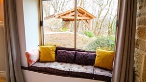 A room with a built-in bench by a large window, a bed with a floral bedspread, and a stone wall. - Gîtes de France® - La Bouyssounade (CRAYSSAC)