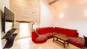A modern living room with a stone wall, a large sectional sofa with red cushions, a wooden coffee table, and a flat-screen TV mounted on the wall. - Gîtes de France® - La Bouyssounade (CRAYSSAC)