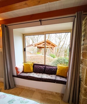 A room with a built-in bench by a large window, a bed with a floral bedspread, and a stone wall. - Gîtes de France® - La Bouyssounade (CRAYSSAC)