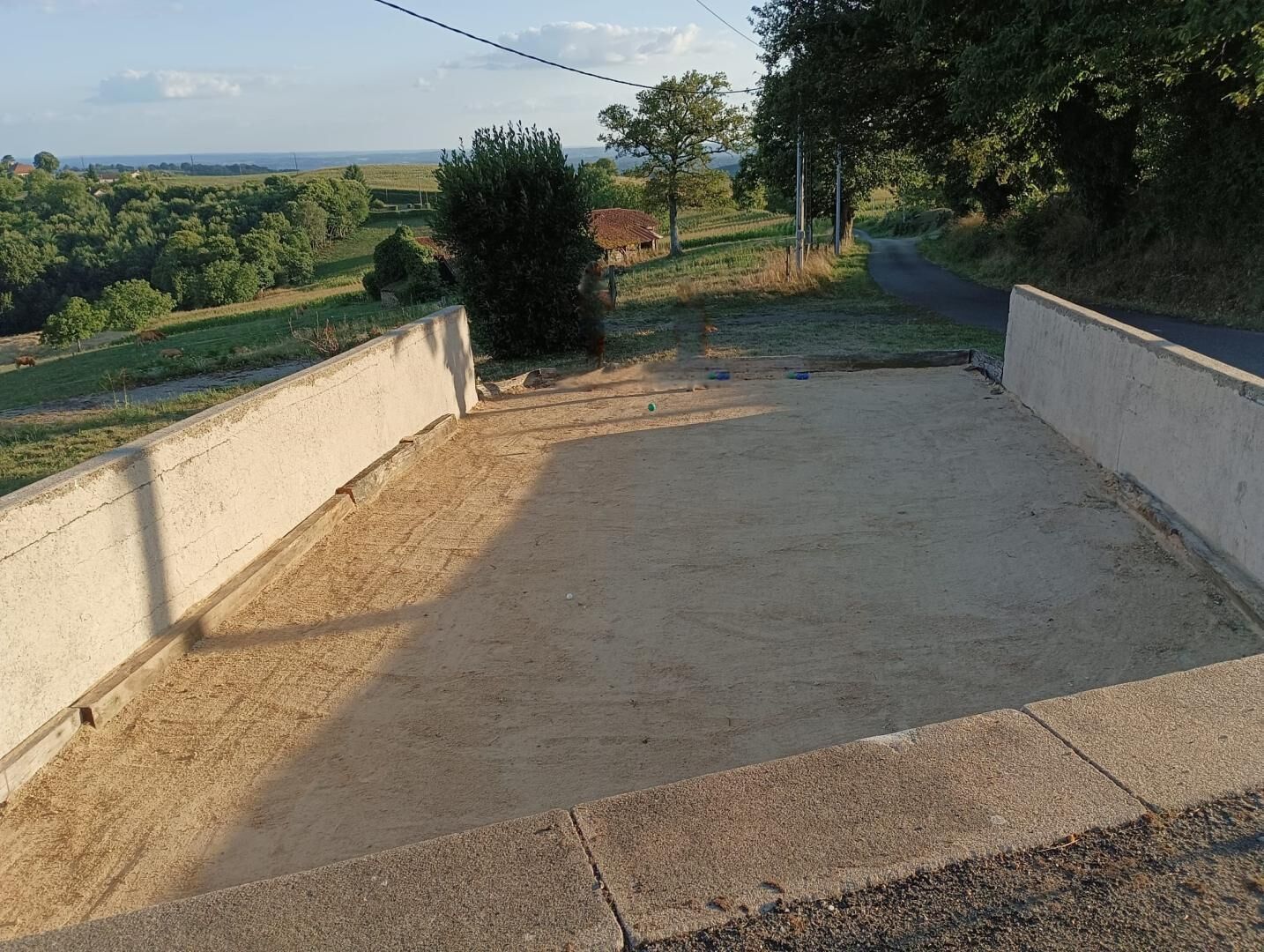 A concrete viewing platform with a dirt floor, surrounded by a low wall, overlooking a rural landscape with fields and a distant house.