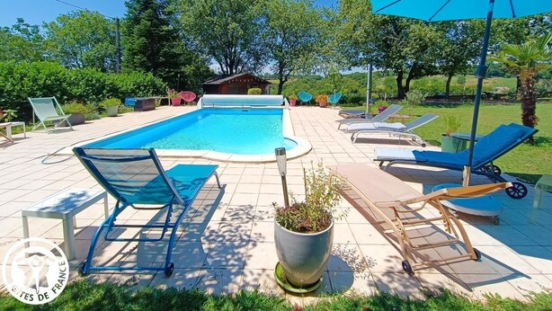 A rectangular swimming pool with lounge chairs and umbrellas, surrounded by greenery and a small building in the background.
