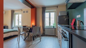 Habitación de hotel compacta con cama, una mesa de comedor con sillas, kitchenette y ventana con cortinas.