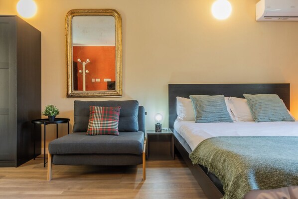 A hotel room with a sofa, a bed with multiple pillows, a nightstand with a lamp, a mirror, and an air conditioning unit. - Terrazza Relax a Borgo Dora by Wonderful Italy (Turin)