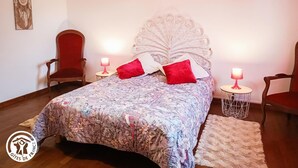 Un dormitorio con colcha estampada, dos almohadas rojas, un cabecero decorativo, dos mesitas de noche con lámparas y una silla roja.